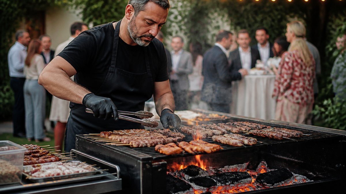 A chef in a black apron grilling skewers and marinated meats over glowing charcoal on a portable grill at an outdoor private event, with guests and festoon lights softly blurred in the background